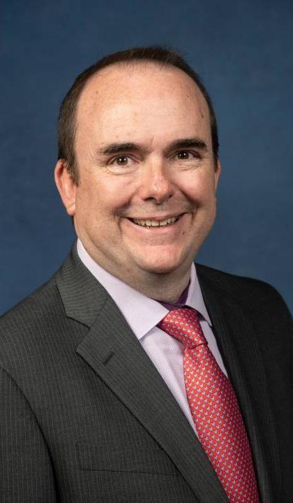 Headshot of Joe Lyons dark suit, light shirt and red tie.